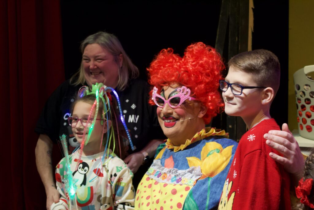 This image shows a family taking part in Angel Eyes NI Touch Tour ahead of our annual Pantomime.  Each year Angel Eyes NI provides the cast of the Pantomime with Touch Tour training. This ensures the cast are aware of vision impairment and are prepared to carry out the touch tour which includes the children and their families coming up to the stage seeing the scenes up close, getting to meet the cast, touch their costumes and hold the props. This year Angel Eyes NI brought 300 people to the pantomime, over 2 showings. The Pantomime is one of Angel Eyes NI and our families highlights of the year!

