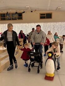 This image shows an Angel Eyes NI family ice-skating together.  This photograph was taken in December 2023 when Angel Eyes NI took 50 family members to an accessible ice-skating experience in Fermanagh.  Each family had the option to take to the ice on ice skates, in their pram or wheelchair, or with the help of an ice buddy - a penguin or a polar to provide extra stability on the ice. This was a wonderful event that allowed the whole family to take part a fun activity together. 